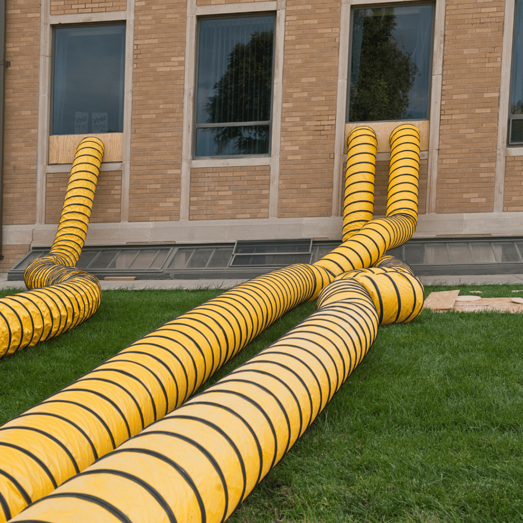 Large yellow and black striped flexible ducts extending from a building's exterior windows down to the grassy ground for ventilation or other industrial purposes.